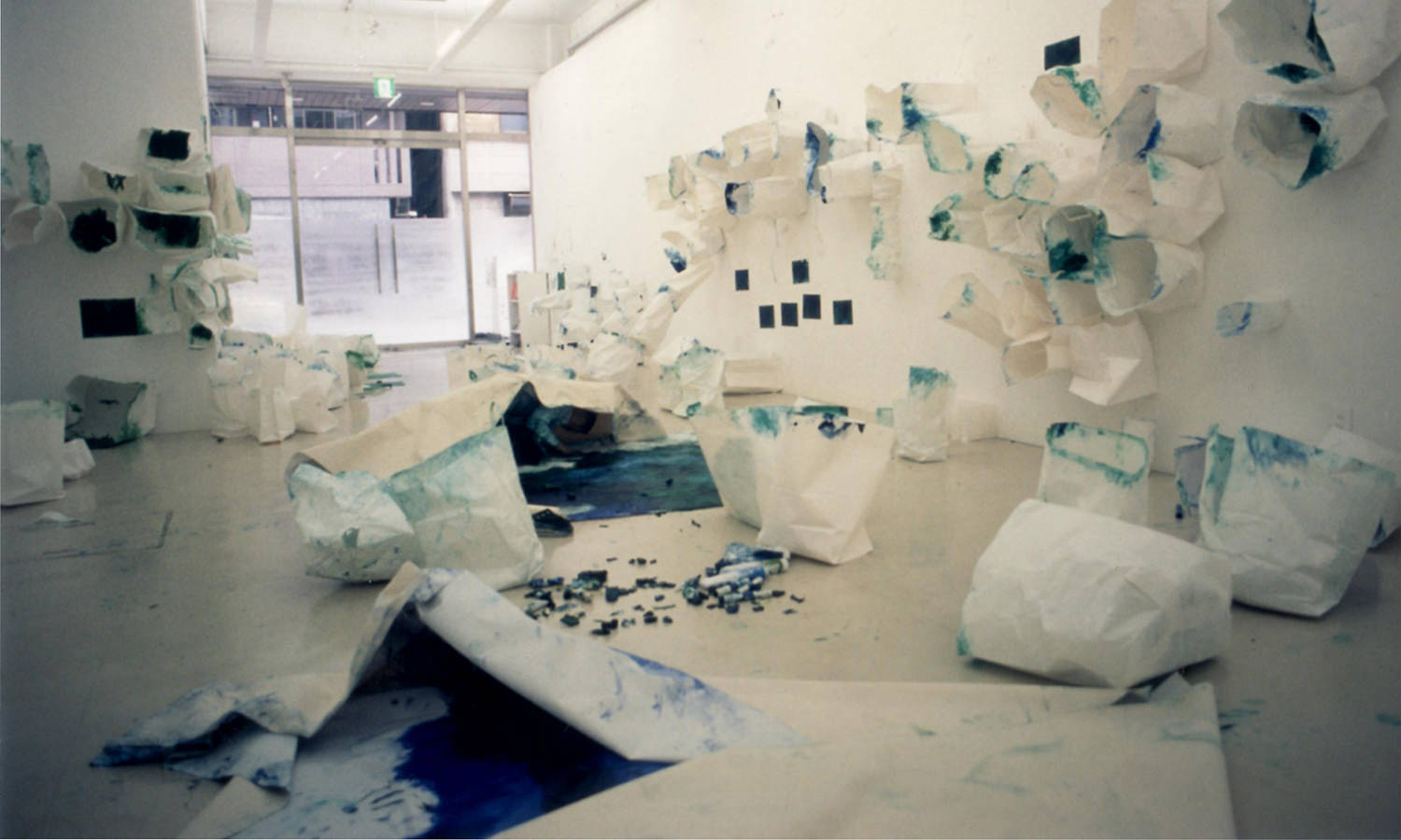 「in the picture/ out of the picture 絵のなか/ 絵のそと｣ 会場制作風景 制作年：2003年／素材：オイルスティック､キャンバス、紙袋、紙／会場サイズ：1390×456.5×310cm ／展示場所：人形町エキジビットスペースVision's（東京）／撮影：柳場大 ＊市販の白い紙袋を使用した。